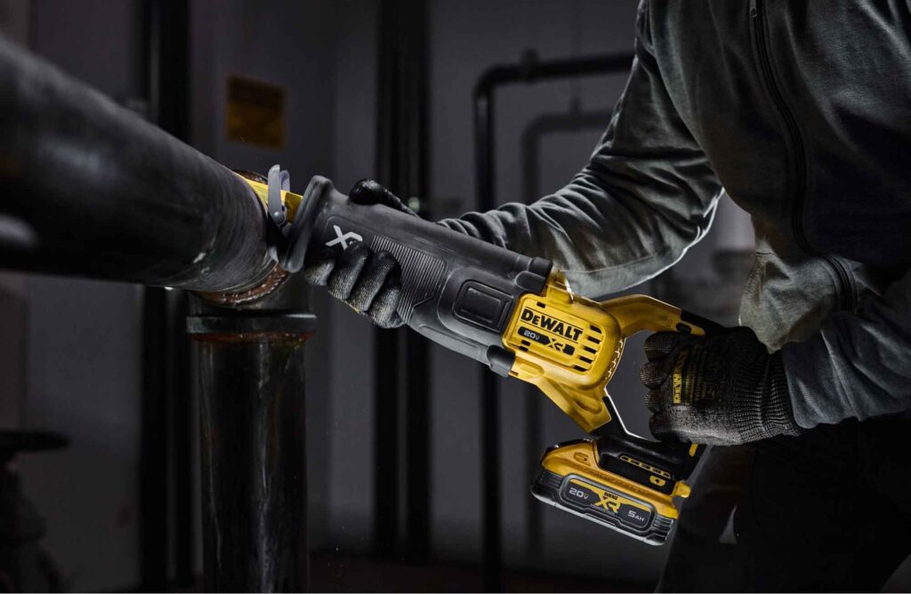 A person wearing gloves uses a DEWALT cordless reciprocating saw to cut through a metal pipe in an industrial setting.
